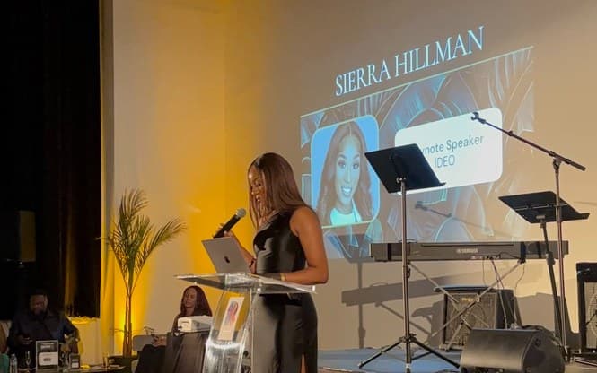 Sierra at a clear lectern on a stage, microphone in hand, her name and Keynote Speaker, IDEO projected behind her.