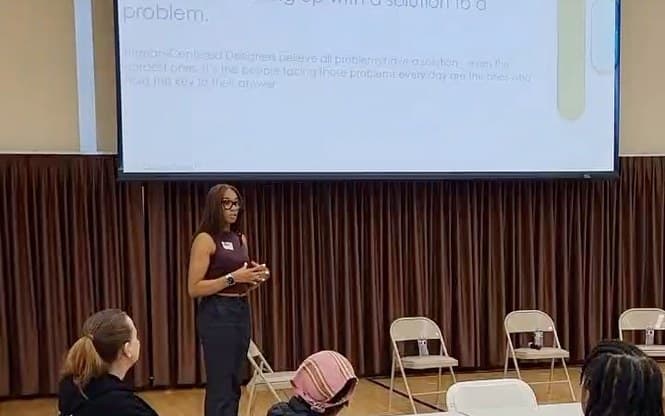 Sierra standing before a slide that defines human-centered design, speaking to a room of people at round tables.